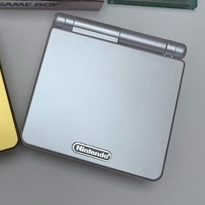 Puede incluir: Una consola de juegos portátil Game Boy Advance SP plateada con un logotipo de Nintendo plateado en la parte delantera.