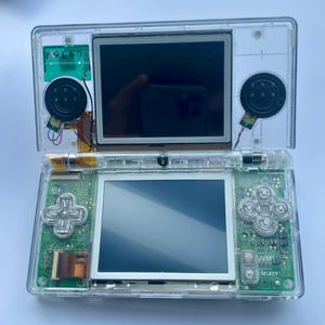 May include: A transparent Nintendo DS handheld game console, open to reveal two screens. The top screen is black, and the bottom screen is silver. The console has a directional pad, buttons, and speakers. The internal components are visible.