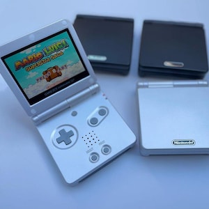 May include: A white Nintendo Game Boy Advance SP console is open, showing the "Mario & Luigi: Superstar Saga" game. Three other Game Boy Advance SP consoles in silver and black are also visible.