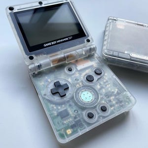 Könnte beinhalten: Eine durchsichtige, transparente Game Boy Advance SP Handheld-Spielkonsole mit schwarzen Knöpfen und einem schwarzen Bildschirm, auf dem der Text "GAME BOY ADVANCE SP" angezeigt wird.