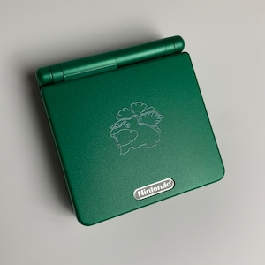 May include: A green Nintendo Game Boy Advance SP handheld console. The console has a silver character emblem and the Nintendo logo. The clamshell design is closed, showing the top surface.