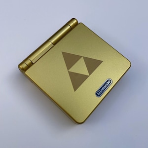 May include: Gold Nintendo Game Boy Advance SP handheld console. The device has a gold finish, a brown Triforce symbol, and a Nintendo logo. The clamshell design is visible in the image.