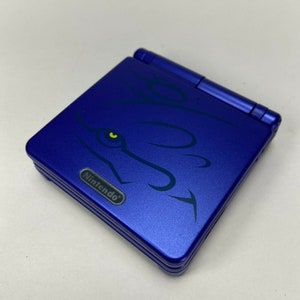May include: A blue Nintendo Game Boy Advance SP handheld game console. The console features a stylized graphic of a wolf's head with a yellow eye. The Nintendo logo is visible on the front.
