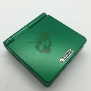 May include: A green Nintendo Game Boy Advance SP handheld console. The device has a dark green graphic on the top cover and a silver Nintendo logo. The console is closed and has a metallic finish.
