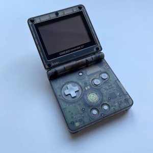 Puede incluir: Una consola de juegos portátil Game Boy Advance SP transparente, abierta e inclinada. La pantalla es negra y los botones y componentes internos de la consola son visibles. El texto "GAME BOY ADVANCE SP" está impreso en el bisel de la pantalla.