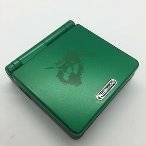 Puede incluir: Una Game Boy Advance SP verde con un logotipo de Nintendo plateado. La consola tiene un diseño verde oscuro con un gráfico estilizado en la parte superior.
