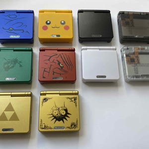 Könnte beinhalten: Eine Sammlung von zehn tragbaren Nintendo Game Boy Advance SP Spielkonsolen in verschiedenen Farben und Designs, darunter blaue, gelbe Pikachu, schwarze, transparente, grüne, rote, weiße und goldene Ausführungen mit spezifischen Grafiken.