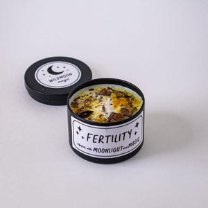 May include: A black metal candle tin with a white label reading FERTILITY infused with MOONLIGHT and MAGIC. The candle wax is yellow and topped with dried herbs and a central wick. The matching black lid features a crescent moon logo.
