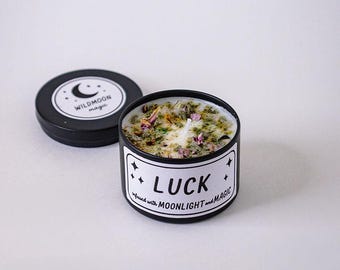 May include: A black metal candle tin with a white label reading LUCK infused with MOONLIGHT and MAGIC. The candle wax is topped with dried herbs and flower petals. A matching black lid with a WILDMOON magic logo sits beside the candle.