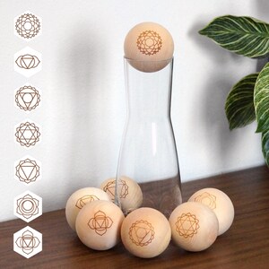 Könnte beinhalten: Eine klare Glaskaraffe mit einem hölzernen Kugelverschluss mit einem geätzten Design. Sieben Holzspären mit geätzten Chakra-Symbolen sind um die Karaffe angeordnet. Sieben weiße sechseckige Aufkleber mit braunen Chakra-Symbolen befinden sich links.