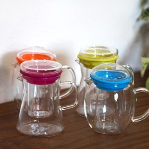 Könnte beinhalten: Vier gläserne Teekannen mit bunten Silikondeckeln. Die Deckel sind rot, orange, gelb und blau. Die Teekannen haben ein eingebautes Sieb.