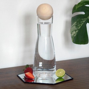 Könnte beinhalten: Eine klare Glasflasche mit einer Holz Kugel, die auf dem Wasser im Inneren schwimmt. Die Flasche steht auf einem sechseckigen Spiegel mit einer Limette und Erdbeeren auf der Oberfläche.