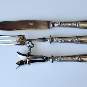 Könnte beinhalten: Dreiteiliges antikes Servierbesteck aus Silber. Enthält ein Tranchiermesser, eine Tranchiergabel und einen Fleischhalter. Die Griffe sind mit floralen Details verziert. Die Messerklinge ist hellbraun.