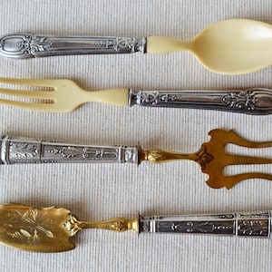 Puede incluir: Cuatro utensilios de servir antiguos: una cuchara, un tenedor y dos tenedores especializados. La cuchara y el tenedor tienen mangos de color marfil y detalles ornamentados de metal plateado. Los otros dos tenedores son dorados con mangos plateados.