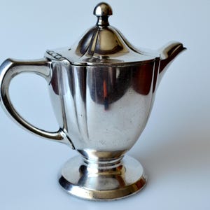 Könnte beinhalten: Eine silberfarbene Teekanne mit Deckel und gebogenem Griff. Die Teekanne hat einen abgerundeten Boden und einen Ausguss. Die Oberfläche der Teekanne ist reflektierend, und der Hintergrund ist weiß.