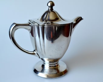 May include: A silver-colored teapot with a lid and a curved handle. The teapot has a rounded base and a spout. The surface of the teapot is reflective, and the background is a plain white.