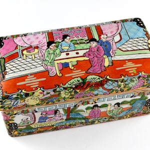 Peut inclure: Boîte décorative rectangulaire peinte à la main, ornée d'une scène détaillée de personnages en costume traditionnel. La boîte présente une palette de couleurs vives de rouge, rose, vert et bleu, avec des motifs floraux et géométriques. Probablement en porcelaine.