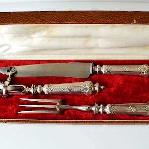 Puede incluir: Un juego de trinchar plateado que consta de un cuchillo, un tenedor para carne y un soporte para carne, todos con mangos ornamentados y en relieve. El set de tres piezas se presenta en un estuche marrón con interior de terciopelo rojo.
