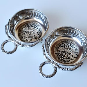 May include: Two silver-colored bowls with ornate detailing. Each bowl has a decorative handle and a detailed emblem in the center. The handles are shaped like a snake. The bowls are on a white surface.