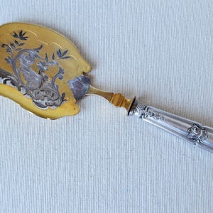 May include: An antique cake server with a gold-colored blade featuring an ornate floral and bird design in silver and black. The handle is silver with decorative detailing and a gold-colored section near the blade.