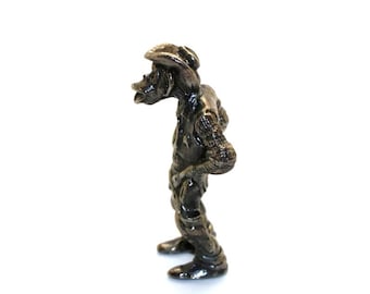 May include: A small, detailed metal figurine of a cowboy, wearing a hat and western attire. The figurine is dark in color and stands on two feet. The cowboy is looking to the left, with his hands behind his back. The figurine is set against a white background.