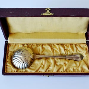 Pode incluir: Uma colher de prata antiga para peneirar açúcar com uma tigela em forma de concha, repousando em uma caixa forrada de veludo bordô com fecho dourado. A colher é exibida sobre uma cama de tecido dourado.