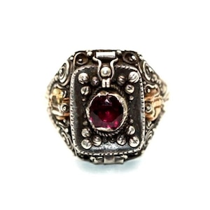 Puede incluir: Un anillo de plata vintage con una piedra preciosa rectangular de color rojo oscuro en el centro. El anillo presenta detalles intrincados, que incluyen pequeñas cuentas de plata y detalles dorados en los lados, sobre un fondo blanco.