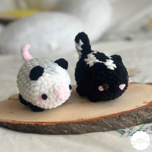 Op de afbeelding: Twee gehaakte knuffels, een zwart-witte skunk en een wit-grijze opossum, zitten op een houten schijf. Beide dieren hebben zwarte ogen en roze neuzen.