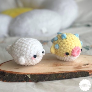 Può includere: Due animali di peluche all'uncinetto, una foca bianca e un pesce palla giallo, sono seduti su una fetta di legno. La foca ha occhi neri e guance rosa. Il pesce palla ha spine blu e gialle e labbra rosa.