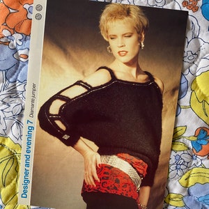 Op de afbeelding: Een zwarte gebreide trui met uitgesneden schouders en een rood-zwarte gestreepte sjaal die om de taille is geknoopt. De tekst "Designer and evening 7 Diana jumper" is gedrukt op de zijkant van de afbeelding.