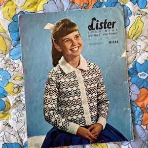 Könnte beinhalten: Ein Vintage-Strickmuster mit einem jungen Mädchen, das einen weißen, roten und schwarzen Pullover mit Muster trägt. Das Muster trägt den Titel "Lister Lochivar Double Knitting" und umfasst die Größe "26 - 28 - 30 ins. chest" und die Nummer "N1414".