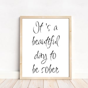 May include: A framed print with the text "It's a beautiful day to be sober" in black script. The print is in a light wood frame and is displayed against a white wall and light wood floor.