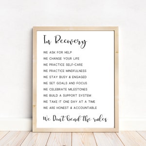 May include: A framed print with the words "In Recovery" at the top, followed by a list of affirmations in black text. The frame is light-colored, and the background is white. The print is displayed on a white wall above a wooden floor.