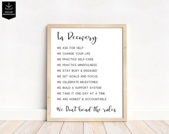 May include: A framed print with the words "In Recovery" at the top, followed by a list of affirmations in black text. The frame is light-colored, and the background is white. The print is displayed on a white wall above a wooden floor.