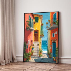 Könnte beinhalten: Gerahmter Kunstdruck, der eine farbenfrohe italienische Straßenszene zeigt. Das Kunstwerk zeigt farbenfrohe Gebäude in Rot, Gelb und Orange, mit Balkonen, die mit Topfpflanzen geschmückt sind. Ein schmaler Kopfsteinpflasterweg führt unter einem strahlend blauen Himmel zum Meer.