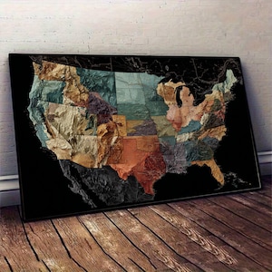 May include: A framed map of the United States, featuring a textured, multi-colored design. The map is set against a black background and is displayed on a wooden floor. The states are represented in various earthy tones.