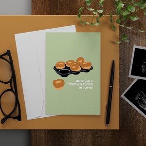 May include: A green card with a cartoon illustration of Yorkshire pudding in a baking tray. The text on the card reads "We've got a Yorkshire puddin in t'oven!"