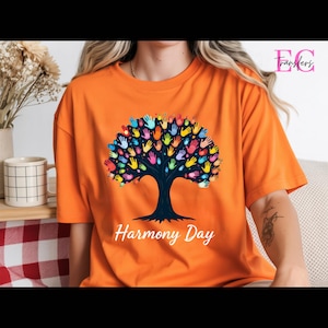 May include: Orange t-shirt featuring a tree design with colorful handprints for leaves. The text "Harmony Day" is printed below the tree in white. The shirt is made of a soft material.