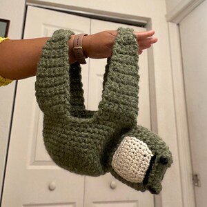 Puede incluir: Una bolsa de mano de crochet verde en forma de perezoso con una cara de crochet blanco y ojos negros.