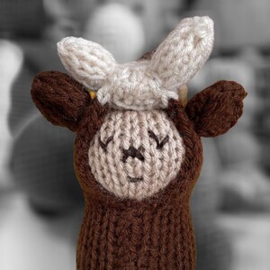 May include: A brown knitted toy cow with white ears and a white face. The cow has embroidered eyes and a smiling mouth.