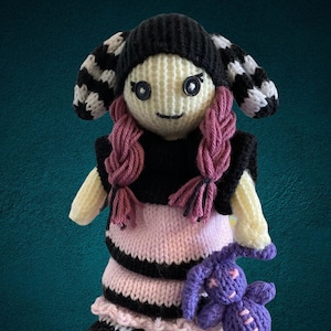 May include: A knitted doll with black and white striped leggings, a pink and black striped skirt, a black sweater, and pink pigtails. The doll is holding a purple knitted bunny.