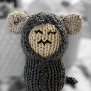 May include: A handmade knitted toy in the shape of a mouse. The mouse has a gray hood and body, with a cream-colored face and ears. The face features embroidered eyes and a mouth. The toy has a small tail.