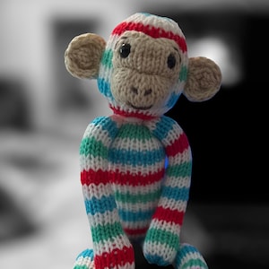 May include: A handmade knitted monkey doll with a striped pattern in red, white, blue, and green. The monkey has tan ears, a tan face, and black eyes. The doll is soft and plush.