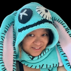 May include: A turquoise blue knitted hooded scarf with pink and white bunny ears. The hood has a stitched-on face with a black X for a nose, a white eye with a black outline, and a stitched-on mouth.