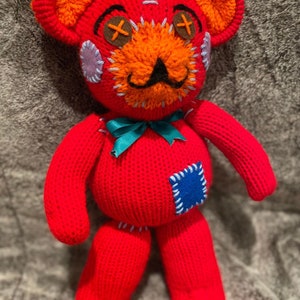 May include: A red knitted teddy bear with orange and blue accents. The bear has a stitched-on face with X-shaped eyes, a black mustache, and a blue patch on its side.