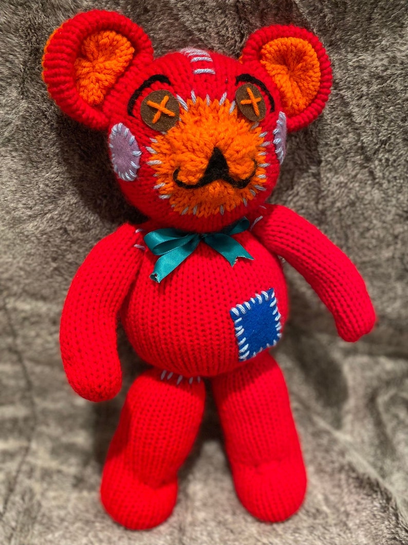 May include: A red knitted teddy bear with orange and blue accents. The bear has a stitched-on face with X-shaped eyes, a black mustache, and a blue patch on its side.