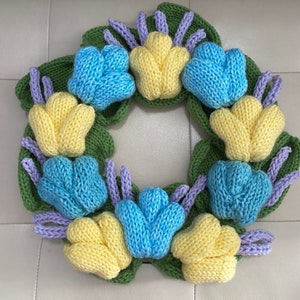 May include: A knitted wreath with yellow, blue, and purple flowers. The wreath is made of green leaves and has a rustic look.