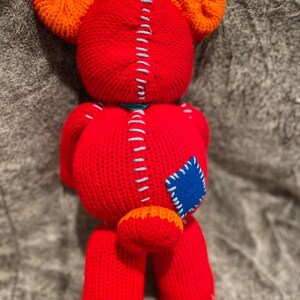 May include: A red knitted teddy bear with orange ears and a blue patch on its side. The bear has a stitched seam down the back and a small orange tail.
