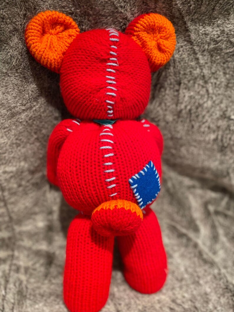 May include: A red knitted teddy bear with orange ears and a blue patch on its side. The bear has a stitched seam down the back and a small orange tail.