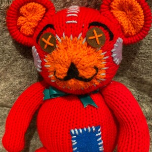 May include: A red knitted teddy bear with orange ears and a stitched-on blue patch. The bear has a stitched-on mustache and X-shaped eyes.